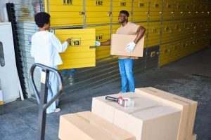 African American man loading a cardboard box into a storage unit, next to a woman manager with a tablet