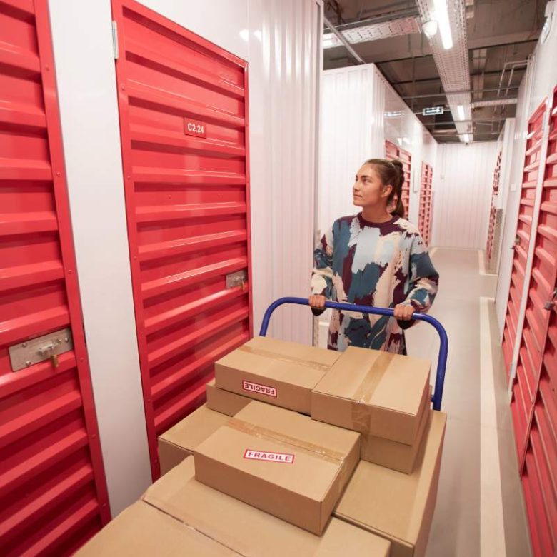 Young woman pushing big cart with many cardboard boxes and looking for door of her storage unit