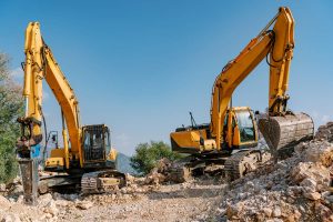 A pair of powerful yellow excavators stands amidst a rocky construction site. The mechanical arms are poised, highlighting the machines' capability for heavy lifting and earthmoving