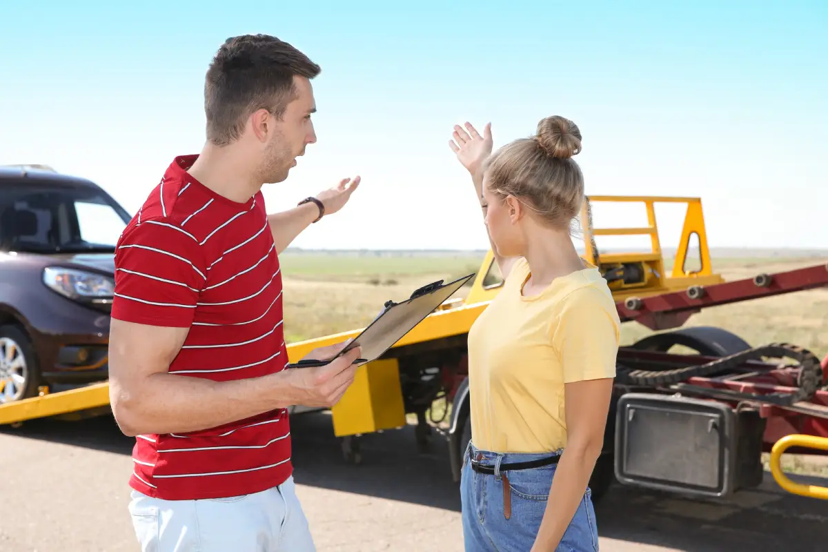 A woman with her hair in a bun wearing a yellow shirt and light blue jeans stands in front of a tow truck